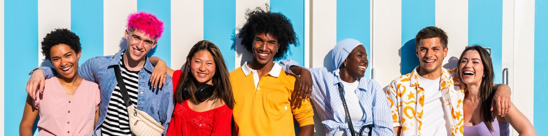 Eine Gruppe junger Menschen steht nebeneinander an einer blau gestreiften Wand. Sie tragen bunte Kleidung und lachen.