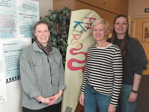Drei Personen stehen vor einer FitKids Beachflag und Ausstellungswand.