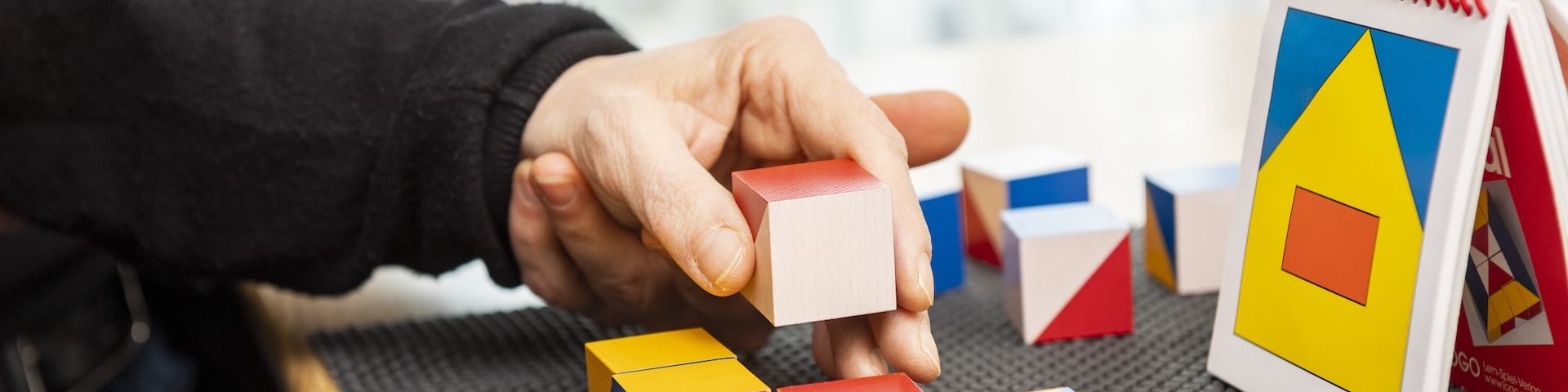 Zwei Hände: eine jüngere Hand unterstützt die ältere dabei, aus bunten Holzklötzen ein vorgegebenes Muster zu legen.